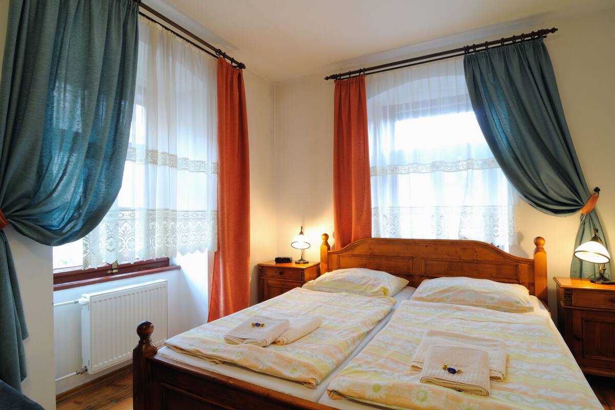 Una cama doble con dos almohadas, un cabecero de madera, una mesita de noche con lámpara y una ventana con cortinas.