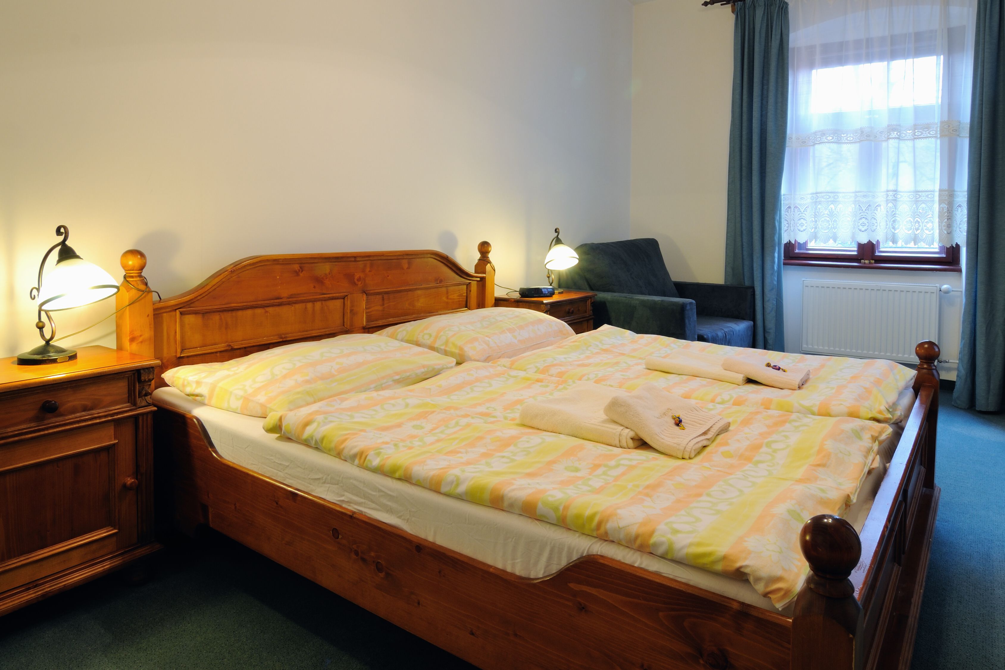 A double bed with a wooden headboard, two bedside tables with lamps, a chair, and a window with curtains.
