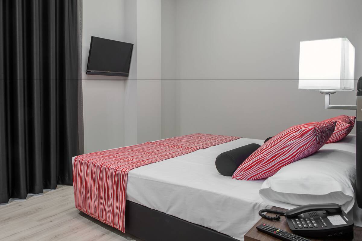 Une chambre d’hôtel moderne équipée d’un lit, d’un téléviseur fixé au mur, d’une table de chevet avec un téléphone et d’une fenêtre avec des rideaux.
