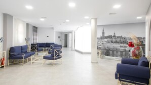 A modern waiting area with blue sofas, a glass coffee table, and a cityscape mural on the wall. - Hotel ITC Artemisa by Soho Boutique (Seville)