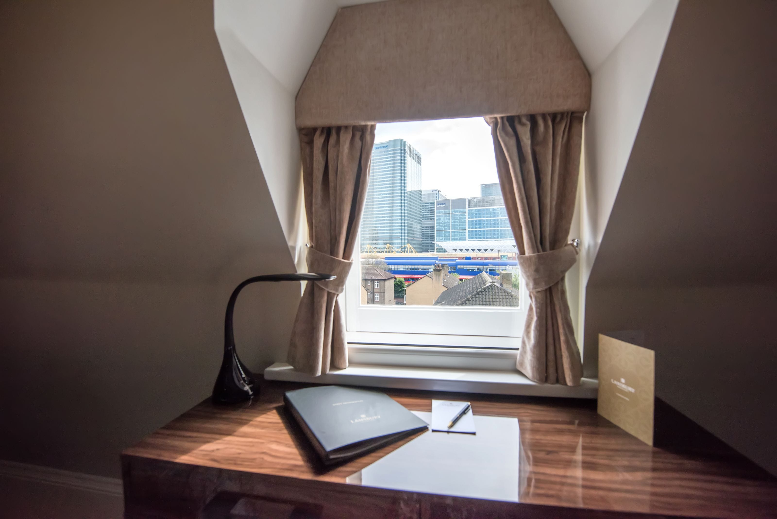 A desk with a lamp, a notebook, and a pen, positioned near a window with curtains, overlooking a cityscape.