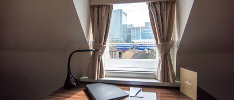 A desk with a lamp, a notebook, and a pen, positioned near a window with curtains, overlooking a cityscape.