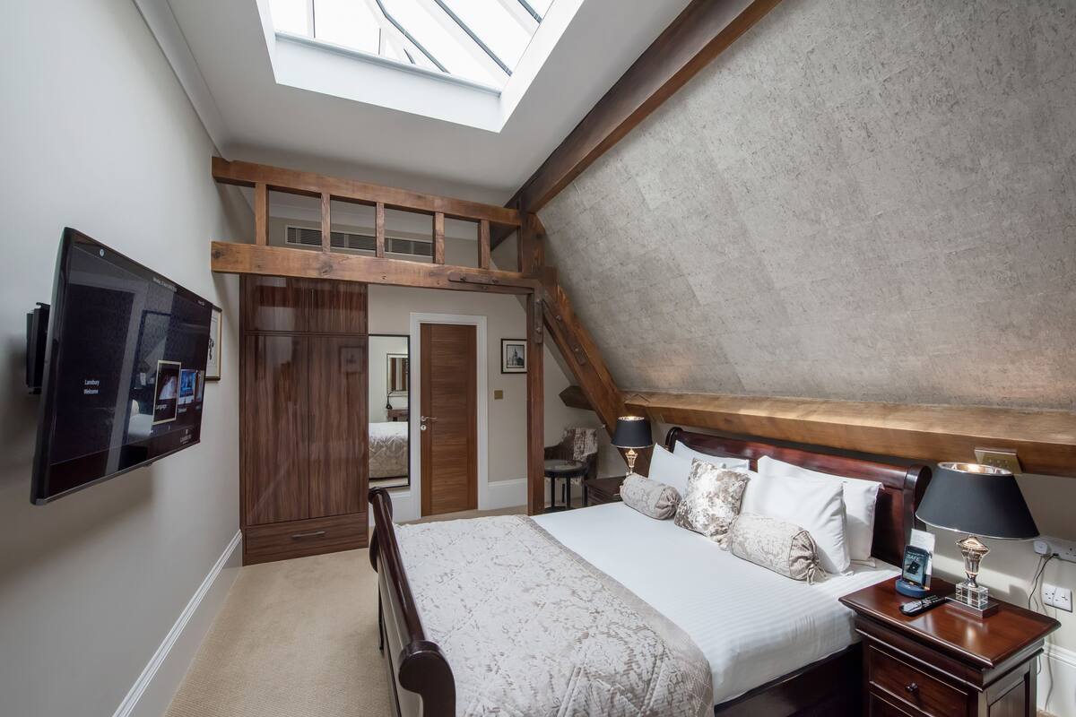 A bedroom with a sloped ceiling, a bed with a patterned bedspread, two bedside lamps, a TV mounted on the wall, and a wooden headboard.