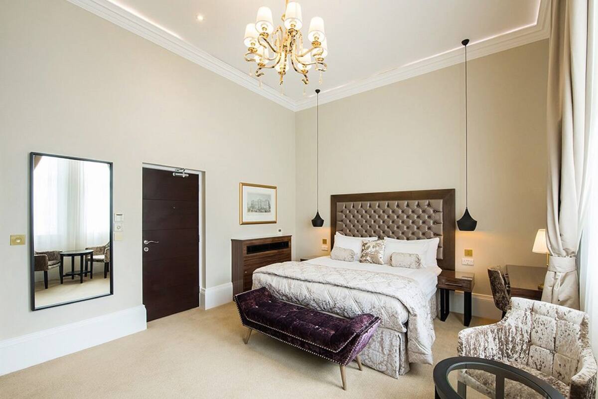 A bedroom with a large bed, a bench, a chair, a dresser, a mirror, and a chandelier.