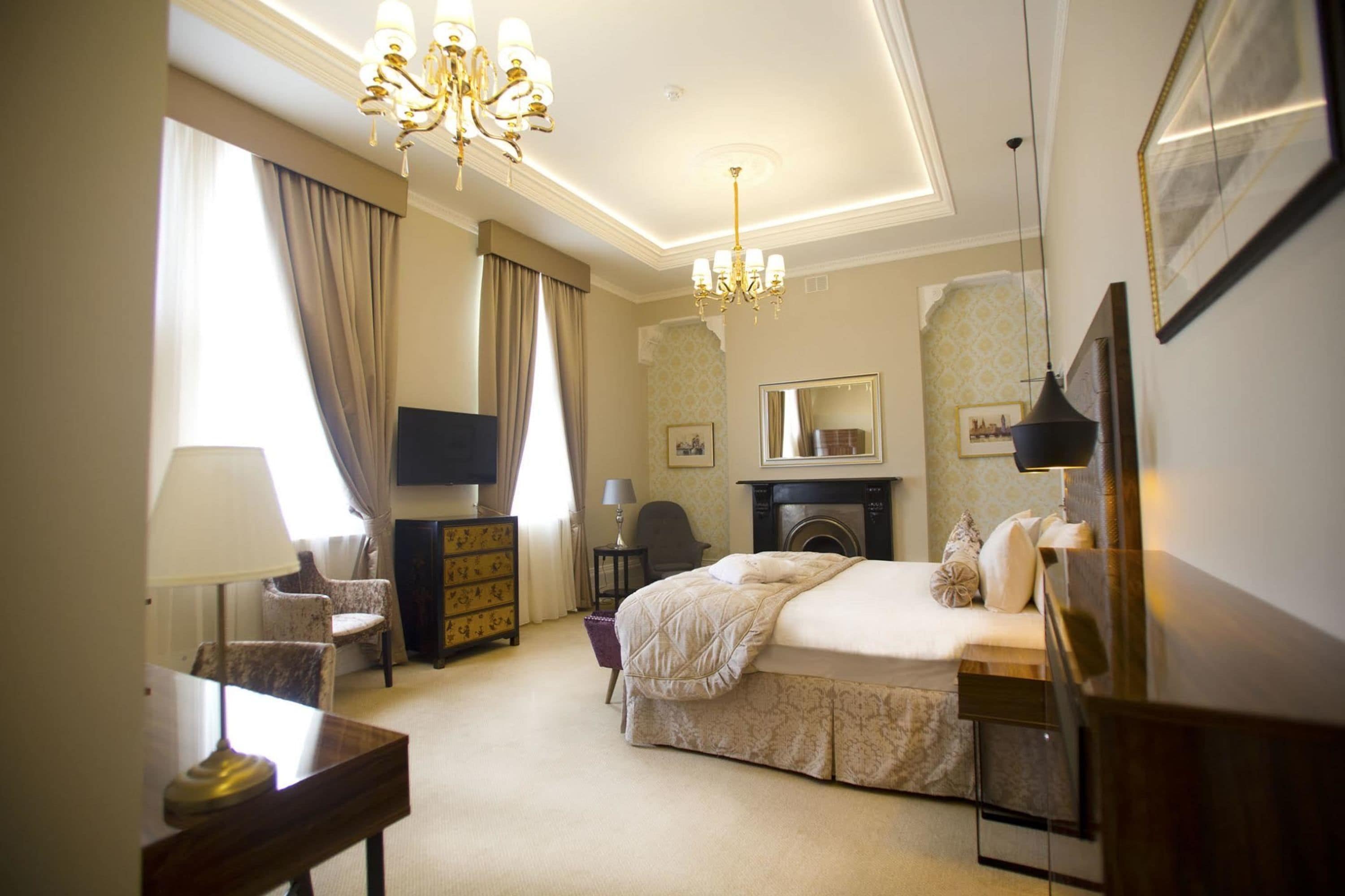 A hotel room with a bed, a television, a desk with a lamp, a chair, a dresser, a fireplace, and a chandelier.