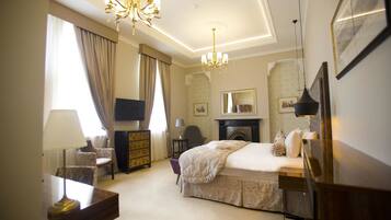 A hotel room with a bed, a television, a desk with a lamp, a chair, a dresser, a fireplace, and a chandelier.