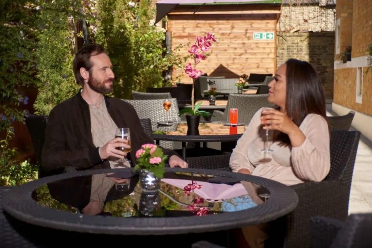 Two people seated at an outdoor table with drinks, surrounded by wicker furniture and potted plants.