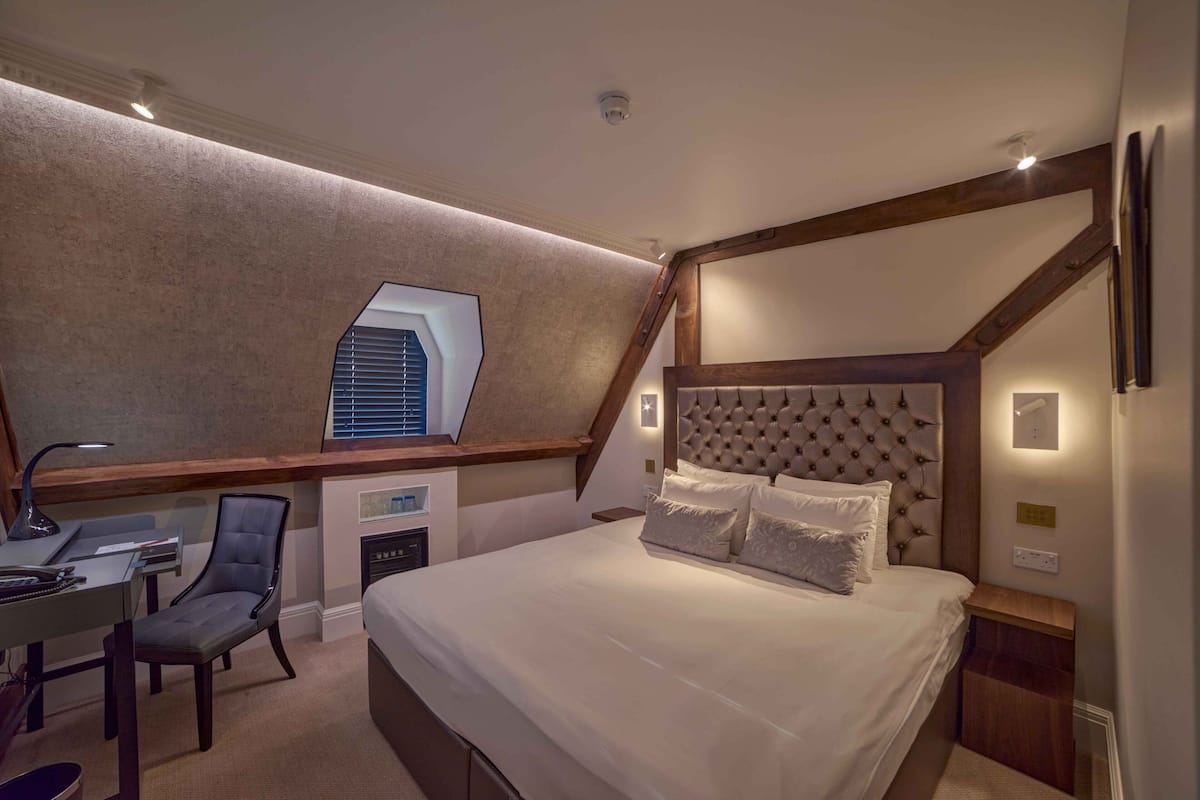A bedroom with a large bed, a desk, and a chair. There is a window with blinds, a headboard, and a small table with a lamp.
