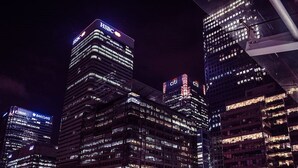 Ein nächtliches Stadtbild mit beleuchteten Wolkenkratzern, darunter erkennbare Firmenlogos wie HSBC und Barclays.