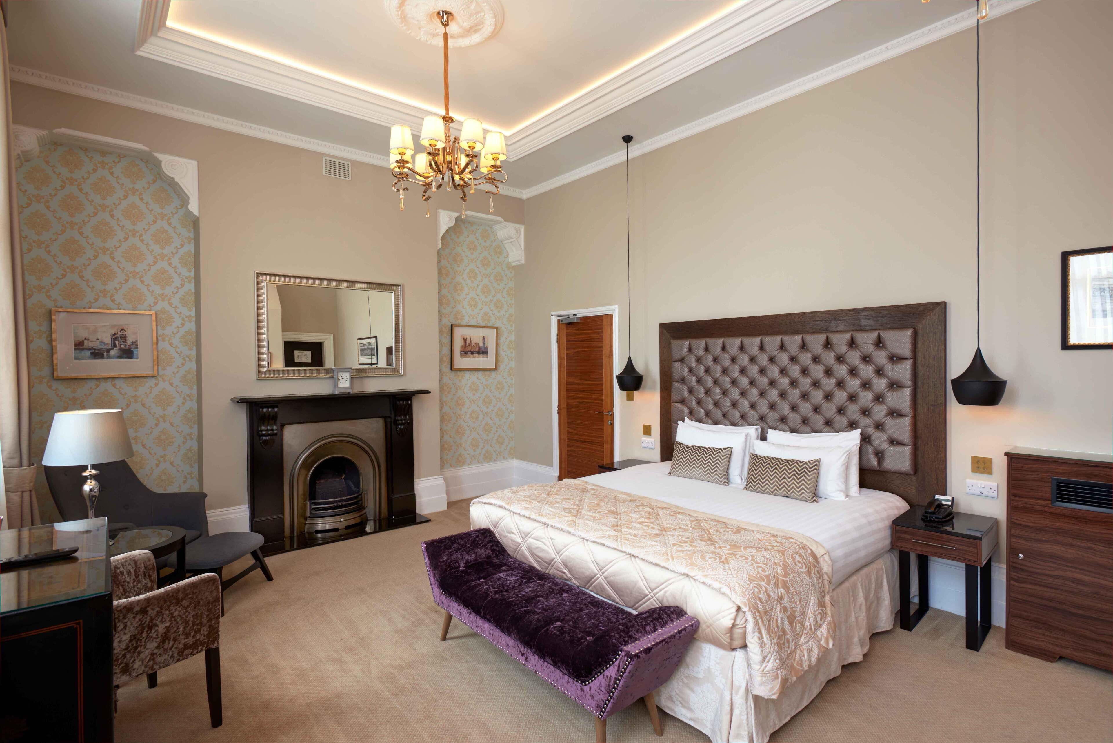 Een stijlvol ingerichte hotelkamer met een groot bed, een open haard, een zithoek met een bank en een bureau met een lamp.