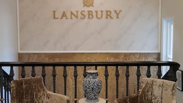 Un área de recepción con una pared de mármol que muestra el nombre "LANSBURY". Hay dos sillones tapizados y un jarrón decorativo sobre una mesita.