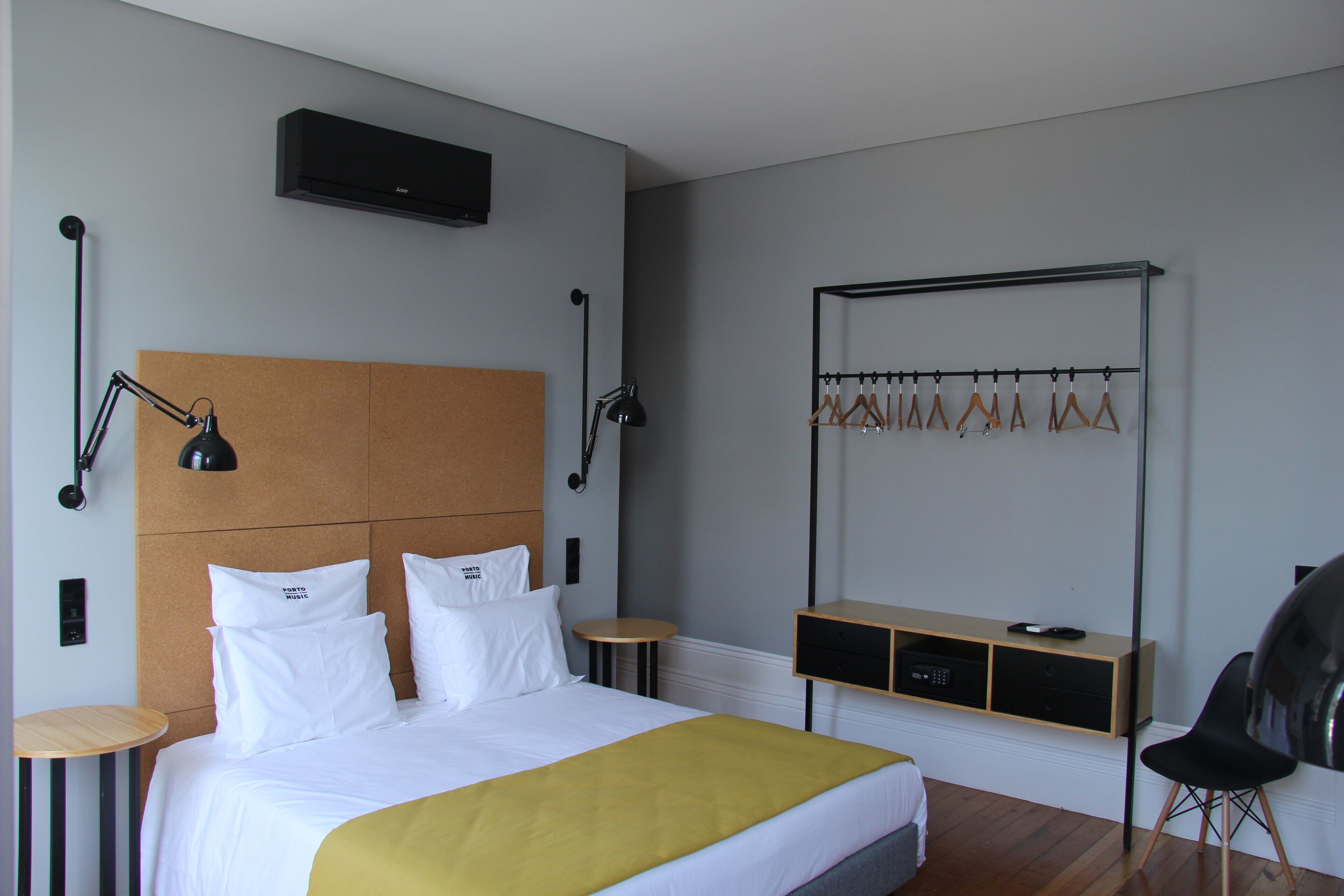 A hotel room with a bed, bedside tables, a desk, a chair, a coat rack, and a wall-mounted air conditioner.