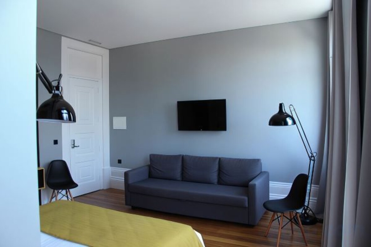 A room with a grey sofa, a TV mounted on the wall, a desk with a chair, and a lamp.