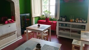 A room with a fireplace, a dining table, chairs, a red bench, a window with a view of greenery, and shelves with dishes and jars. - Flores 105 Guesthouse (Lisbon)