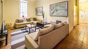 A living room with a sofa, coffee table, and a TV stand. There is a window with a plant on the windowsill. - Flores 105 Guesthouse (Lisbon)