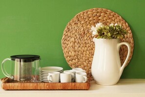 Une table où l’on a préparé du café, avec une cafetière en verre, une tasse blanche et un pichet, le tout posé sur un tapis tissé, devant un mur vert.