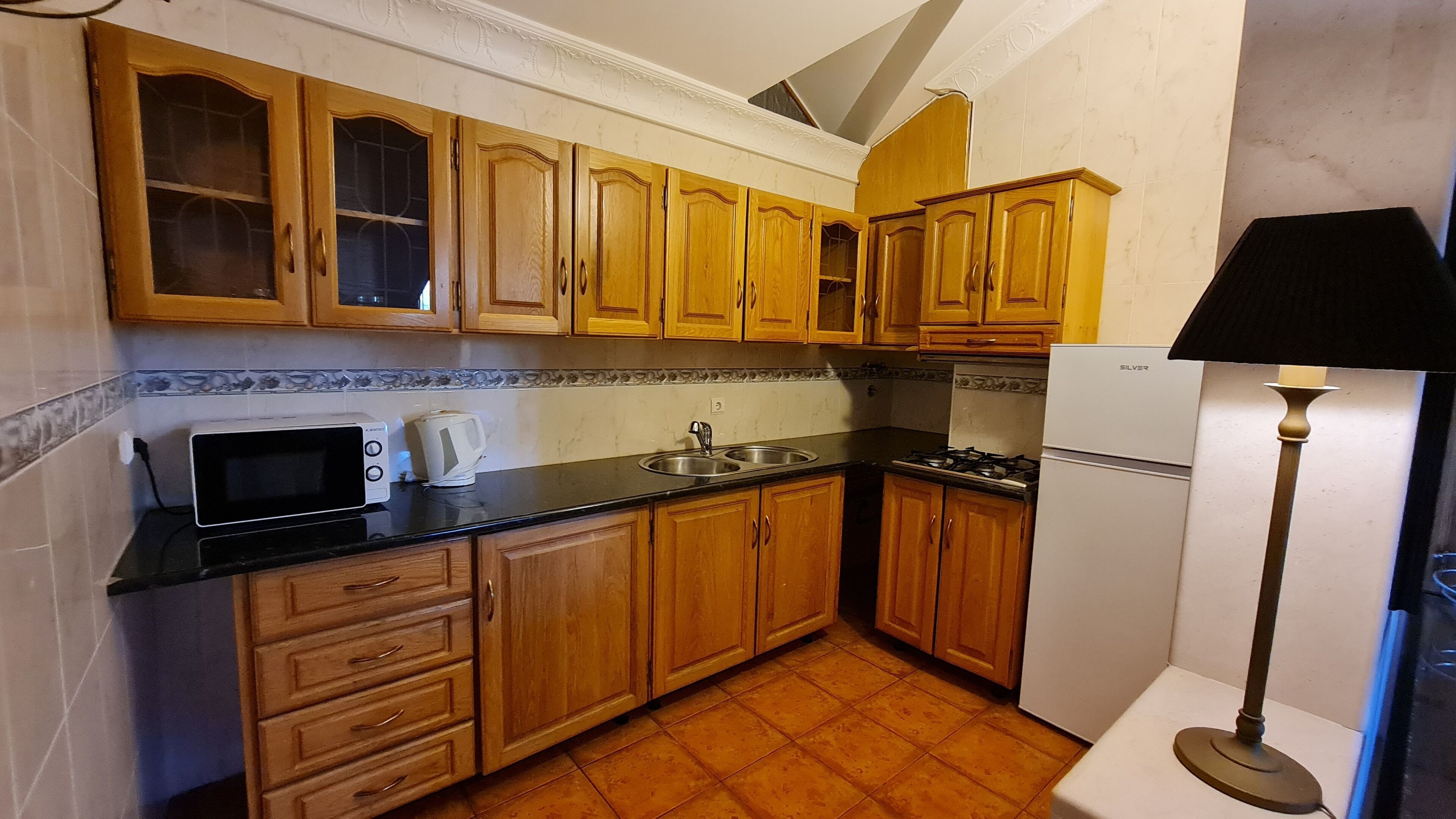 A compact kitchen with wooden cabinets, a microwave, a kettle, a sink, and a refrigerator.