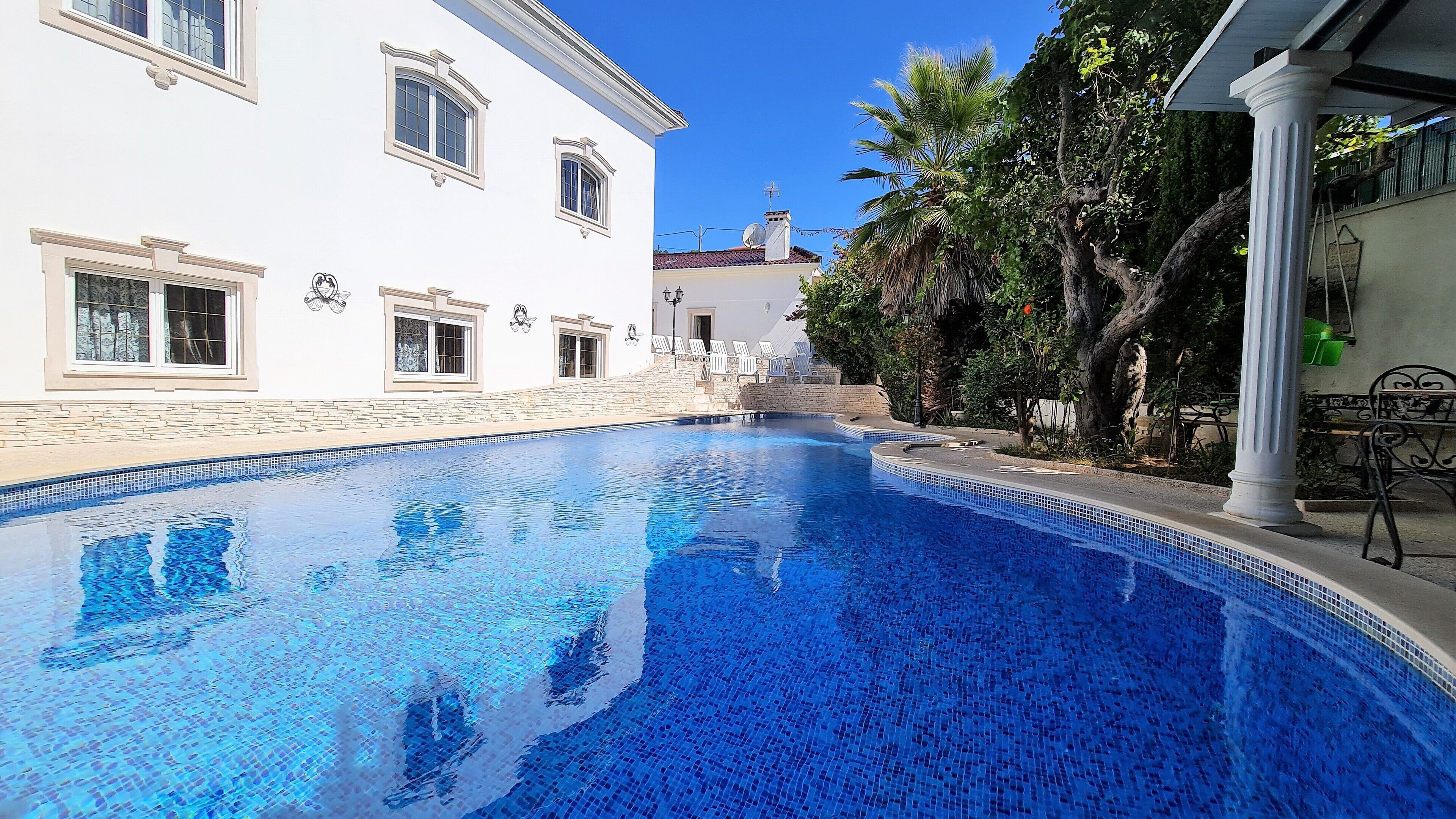 Una piscina con agua azul cristalina, rodeada por un edificio blanco con múltiples ventanas y un muro de piedra.