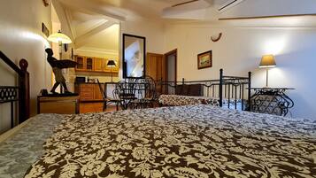 A bedroom with a bed, a chair, a desk, a mirror, and a kitchen area in the background.