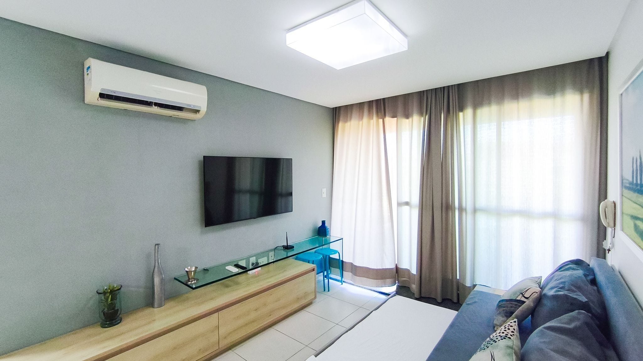Une chambre moderne dotée d’un téléviseur à écran plat, d’un bureau en verre, d’un canapé et d’une grande fenêtre avec des rideaux.