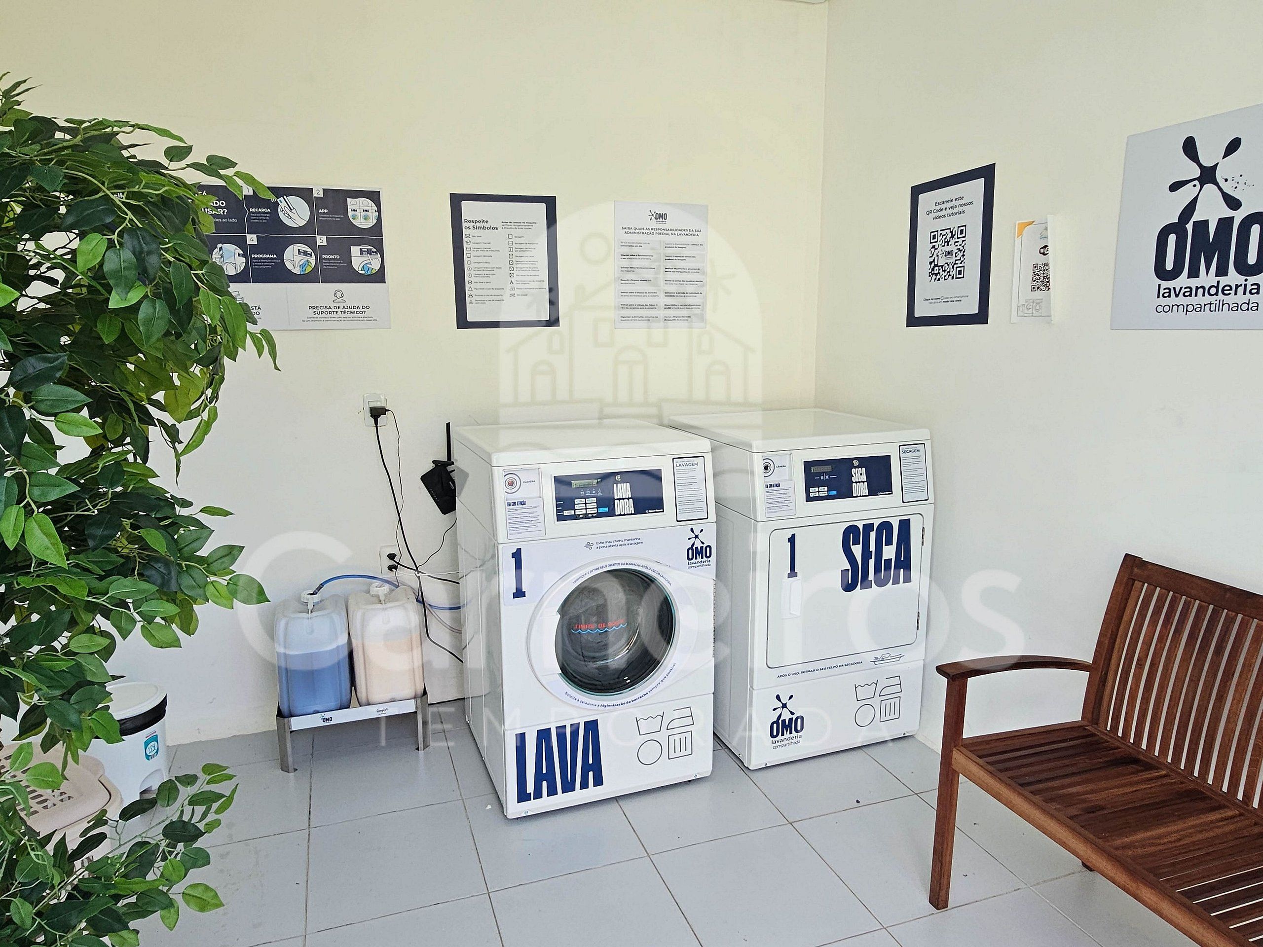 Uma área de lavanderia com duas máquinas de lavar, um banco e cartazes informativos na parede.