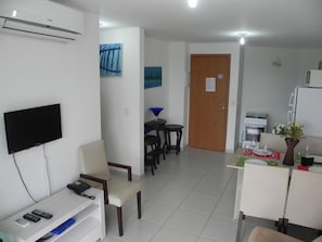 A hotel room with a flat-screen TV, a desk with a phone, a chair, a dining area with a table and chairs, a kitchen area with a refrigerator, and a door with a sign. - Flats no Carneiros Beach Resort (Tamandaré)