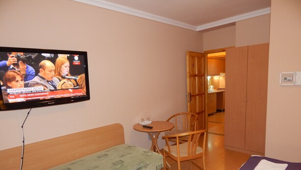 A hotel room with a television displaying a news conference, a green sofa, a wooden chair, a small round table, and a bed with purple bedding. - Hajdu Vendeghaz (Hajduszoboszlo)