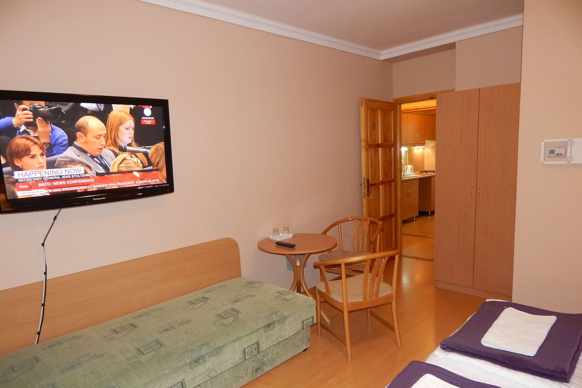 Habitación de hotel con televisor mostrando una rueda de prensa, sofá verde, silla de madera, mesa redonda pequeña y cama con ropa de cama morada.