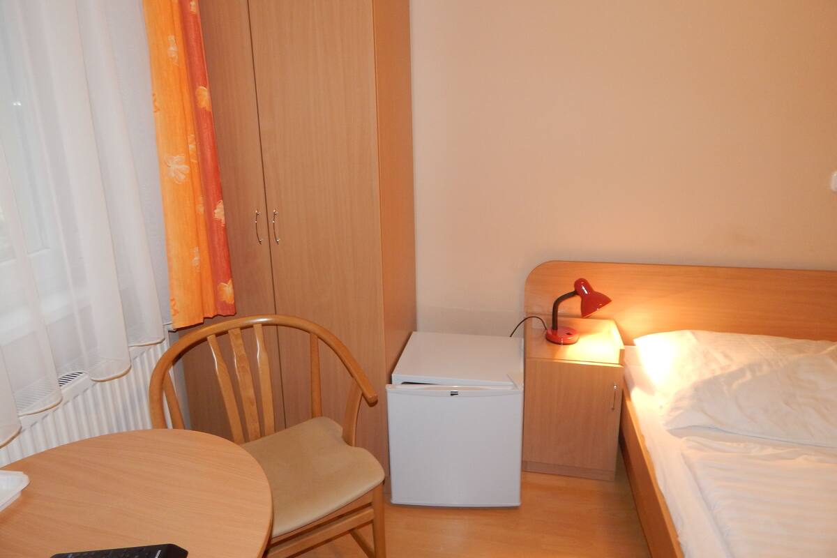 Habitación de hotel compacta con una cama de madera, una mesita de noche pequeña con una lámpara, una silla, un refrigerador compacto y una mesa redonda con un control remoto.