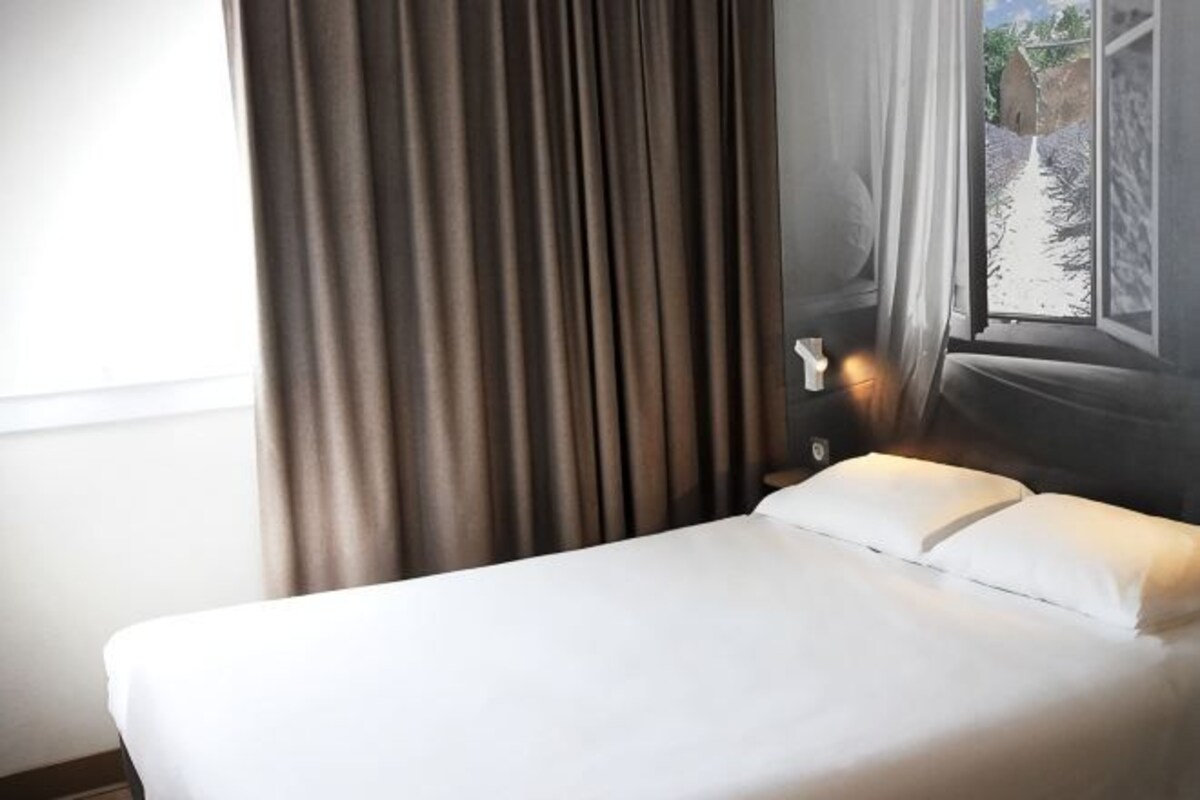 A hotel room with a bed, curtains, a nightstand, and a view of a rocky landscape through a window.