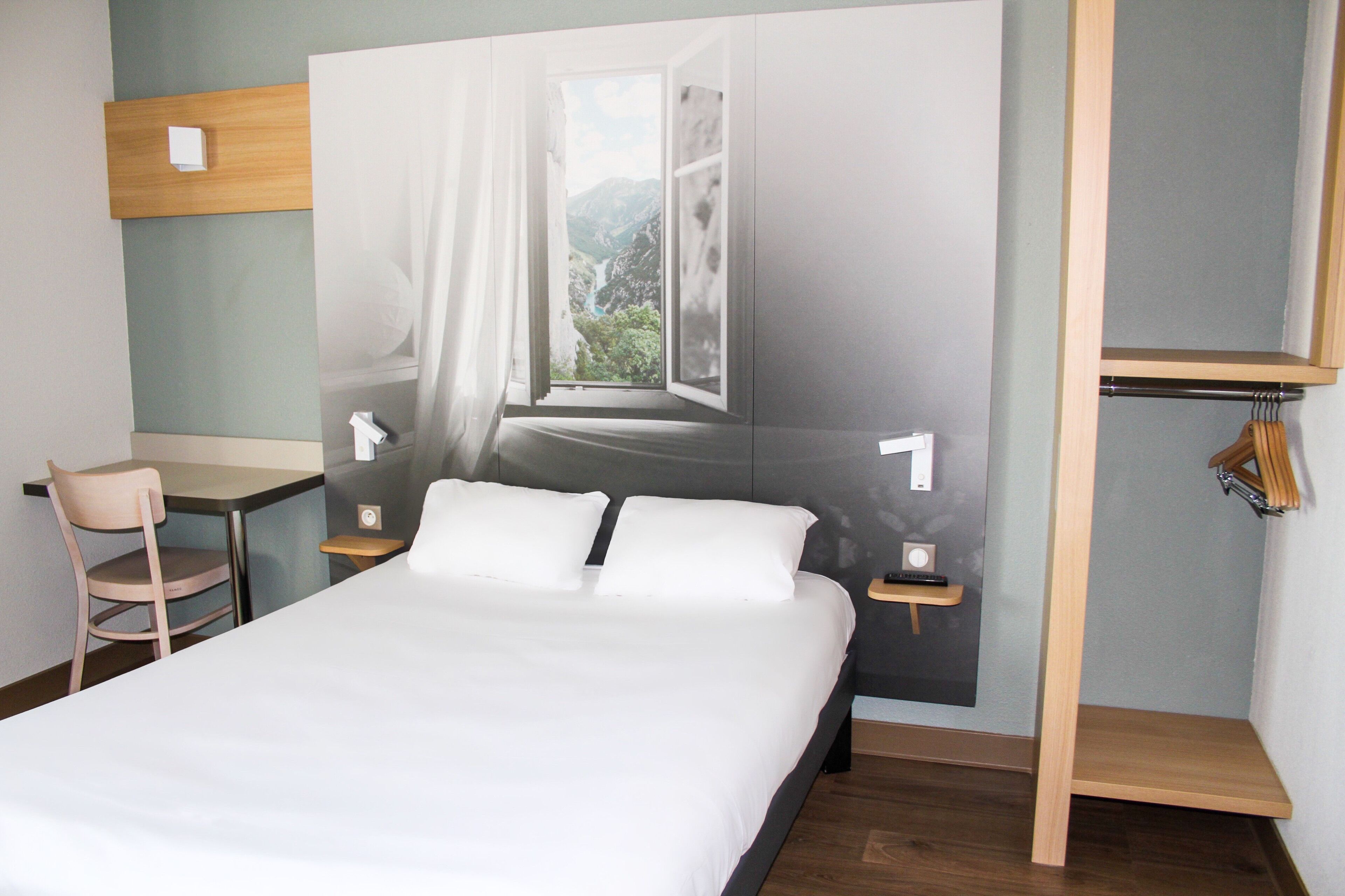 A hotel room with a bed, a desk, a chair, and a view of a mountain through a window.