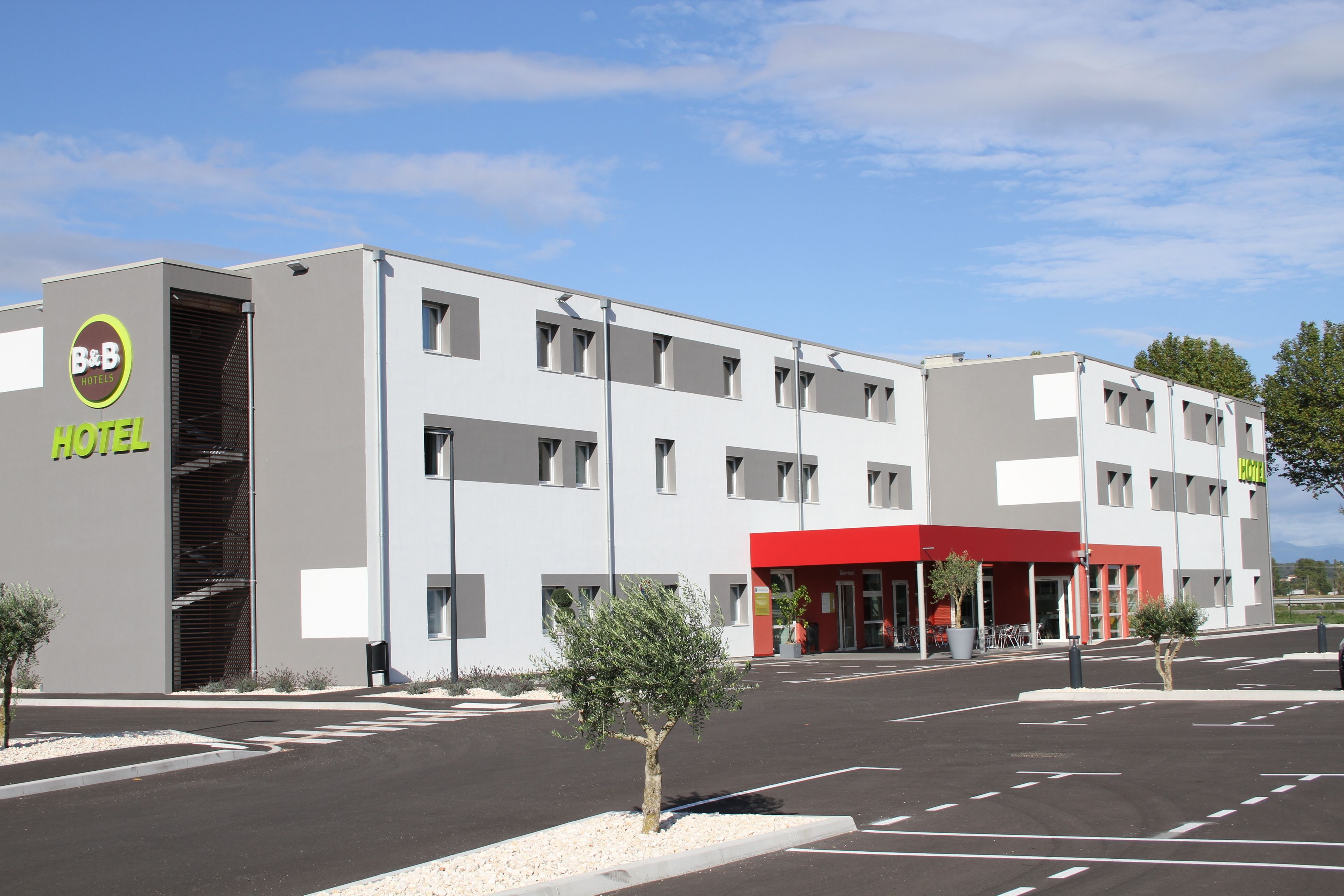 A modern hotel with a red entrance, multiple floors, and a parking lot.