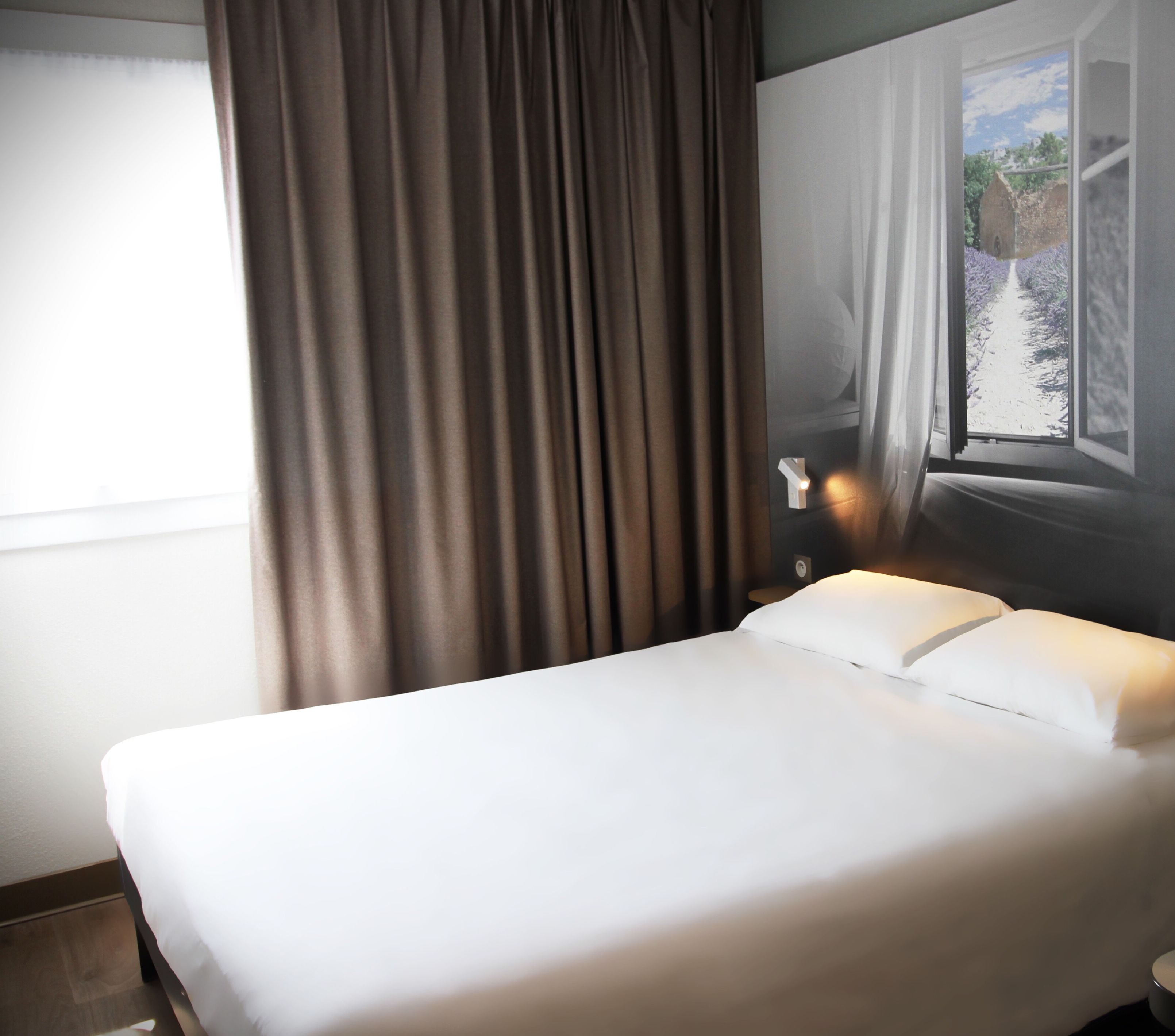 A hotel room with a bed, curtains, a window with a view of a pathway and greenery, and a wall-mounted light.