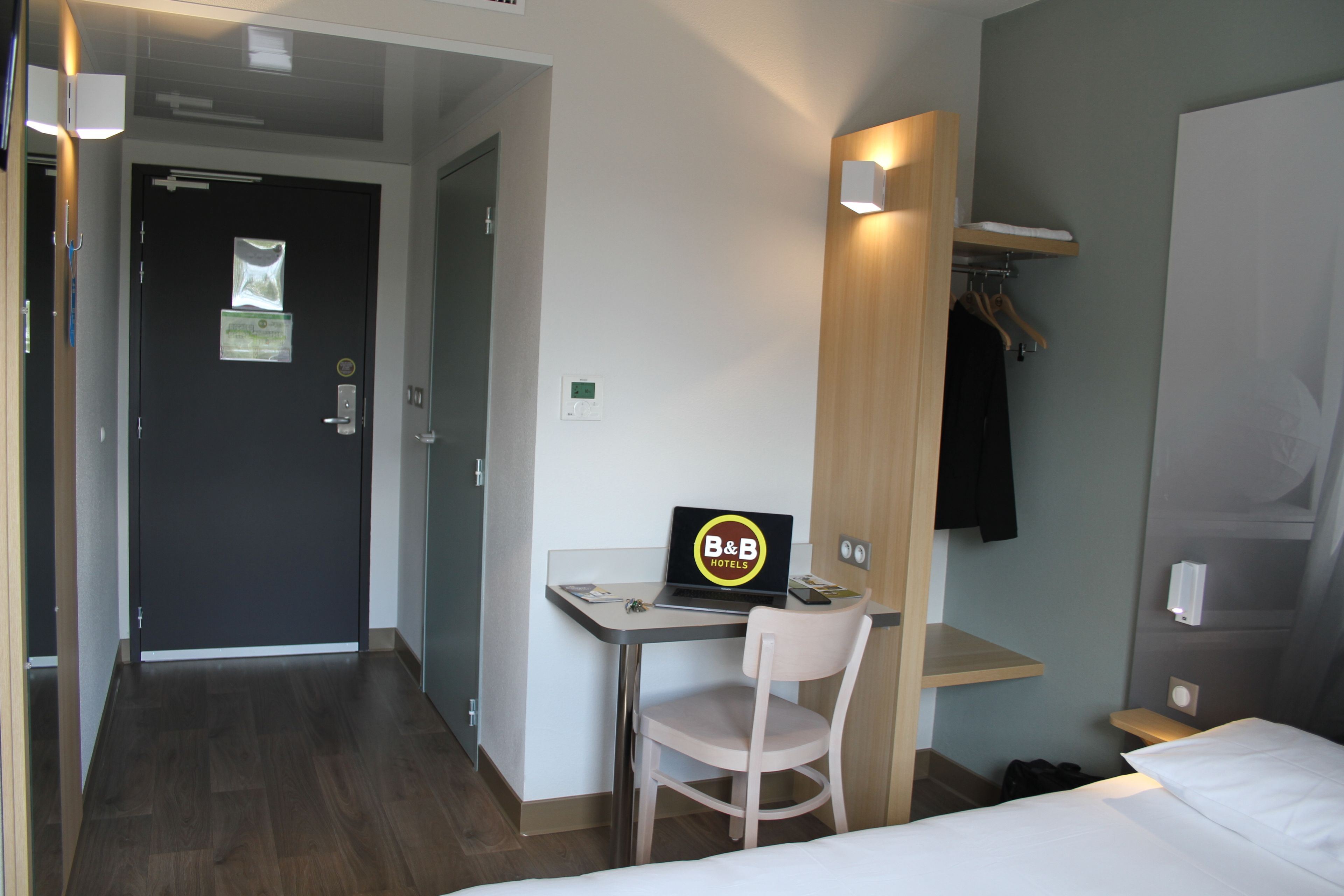 A hotel room with a bed, a desk with a laptop, a chair, a coat rack, and a door.