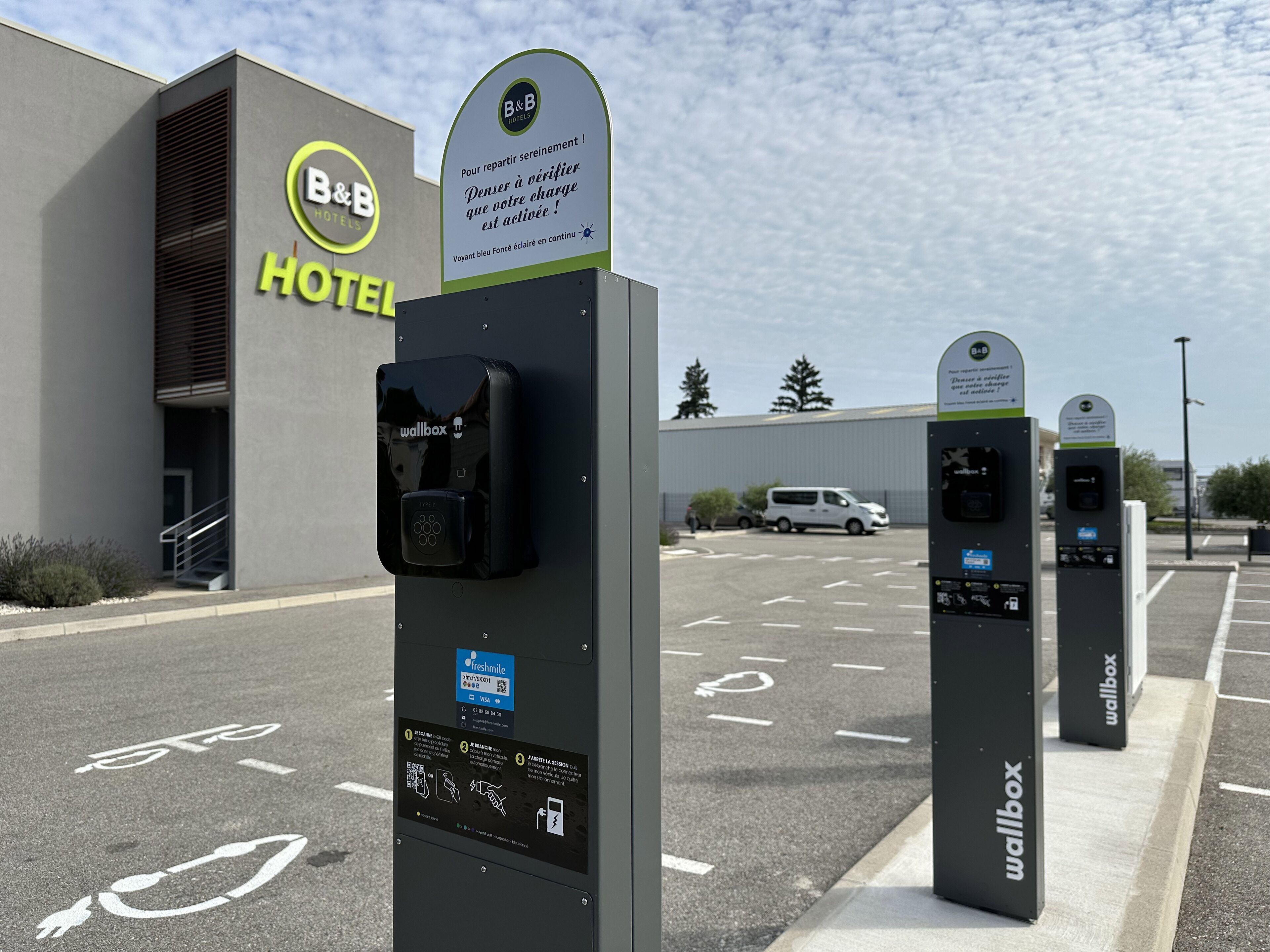 A hotel with a B&B logo, a wallbox charging station, and a parking area with marked spaces.