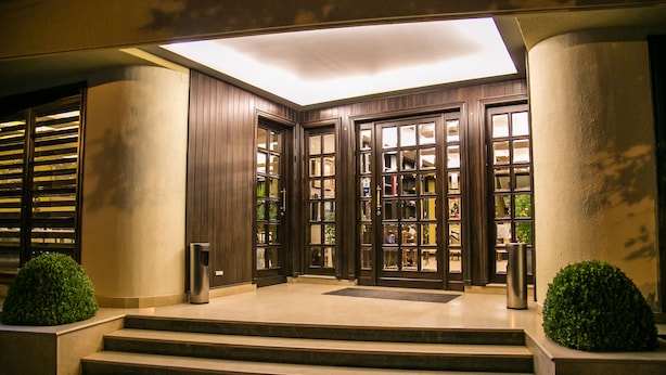 A hotel entrance with a set of steps leading to a door with glass panels, a small trash bin, and two potted plants. - Hotel Victoria (Tirana)