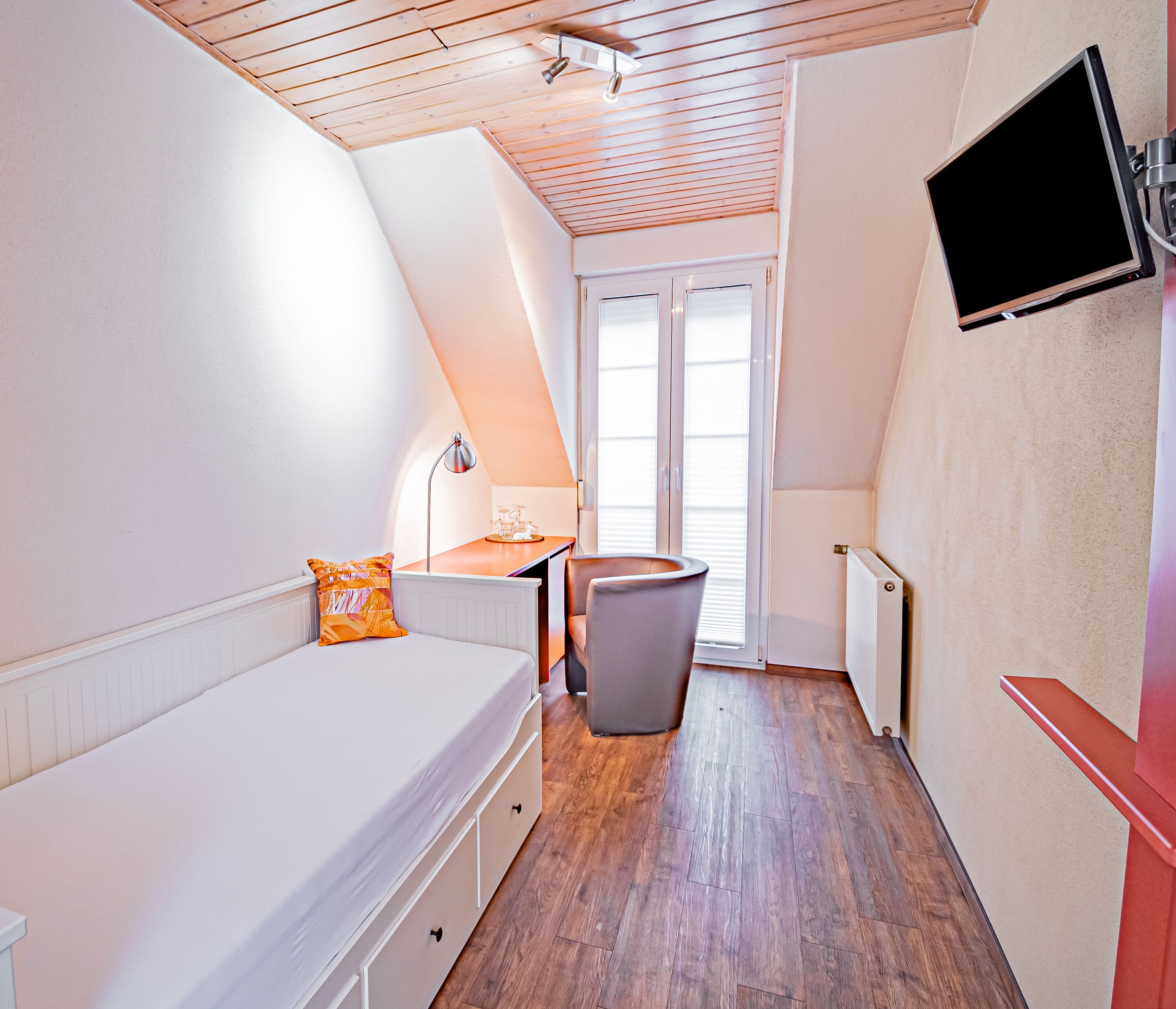 Una habitación estrecha y con techo inclinado, con una cama, un escritorio, una silla y un televisor montado en la pared.