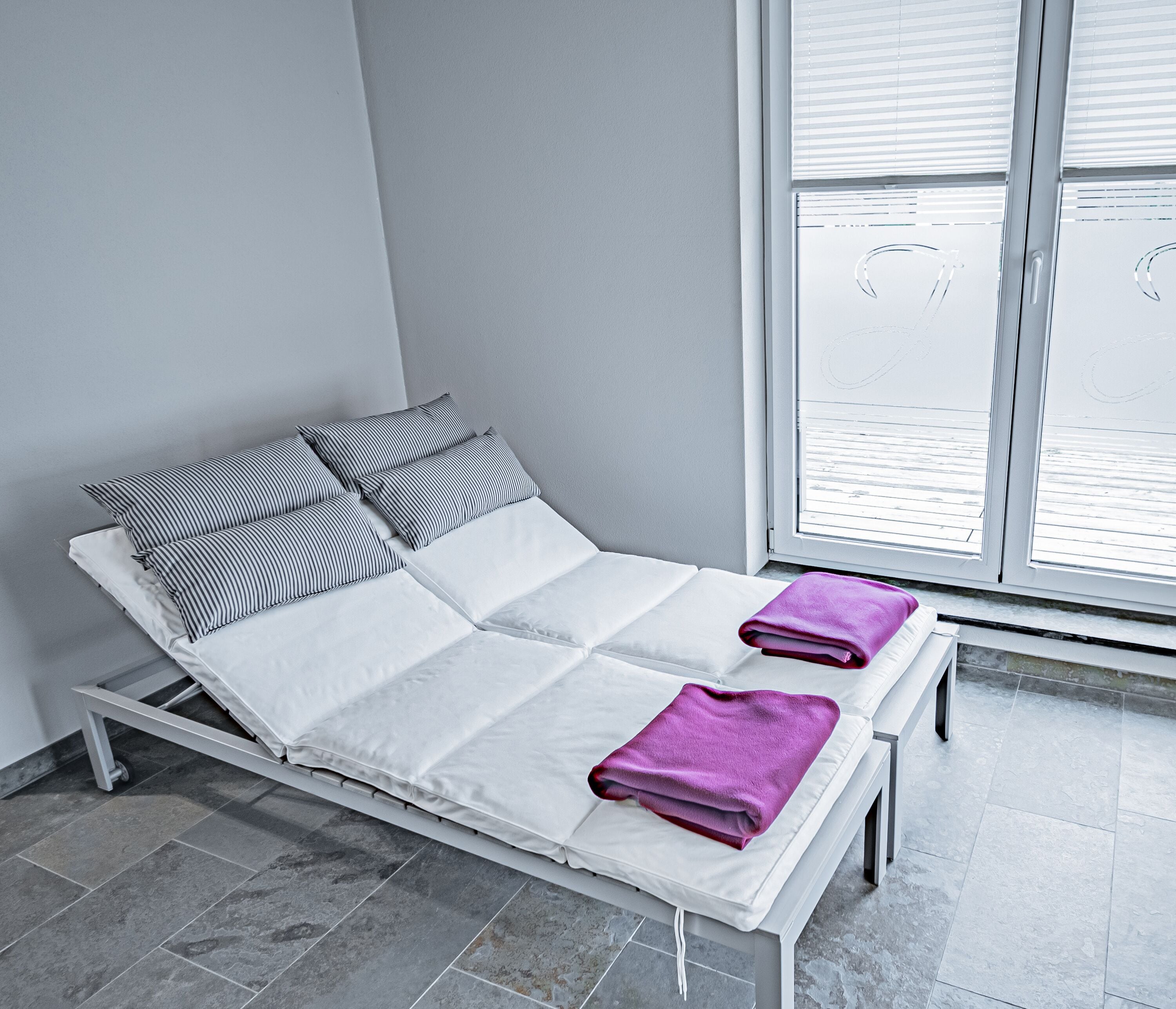 A bed with white bedding and striped pillows, a purple towel, and a window with blinds.