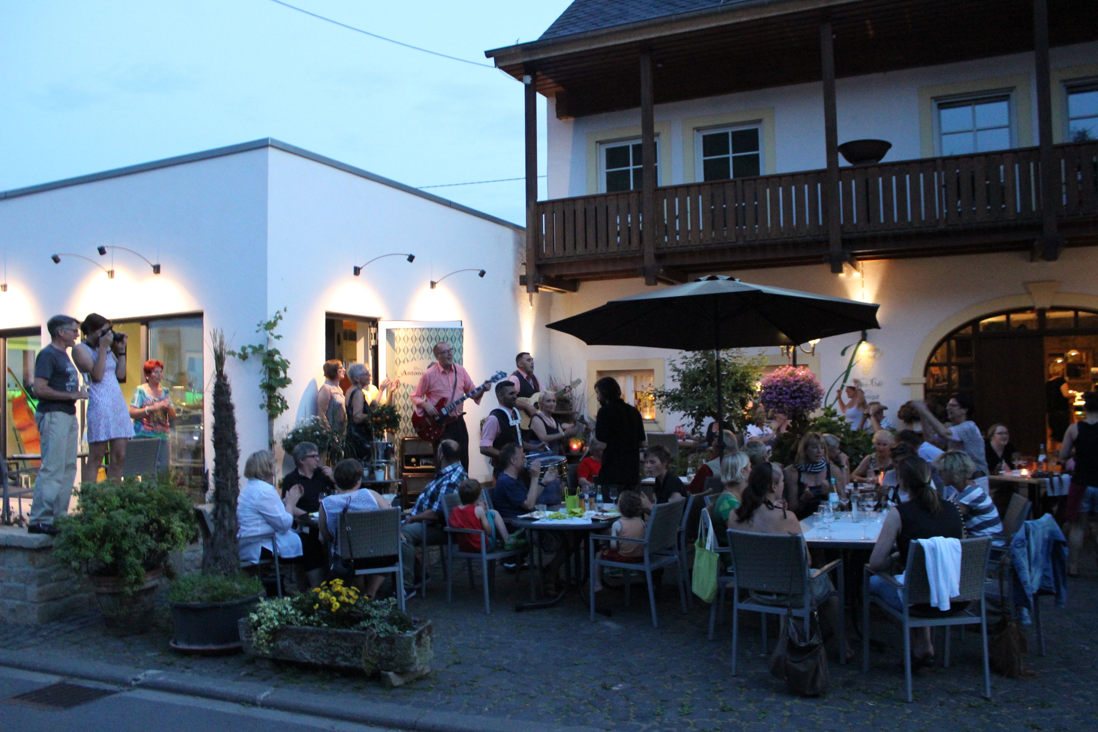 An outdoor dining area with tables and chairs, surrounded by potted plants and a building with a balcony.