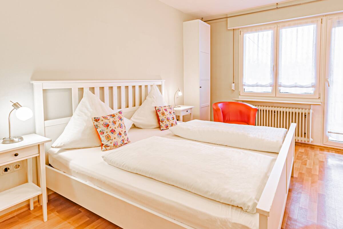 Un lit double avec une literie blanche et des coussins colorés, une table de chevet avec une lampe, une fenêtre avec des rideaux et un radiateur.