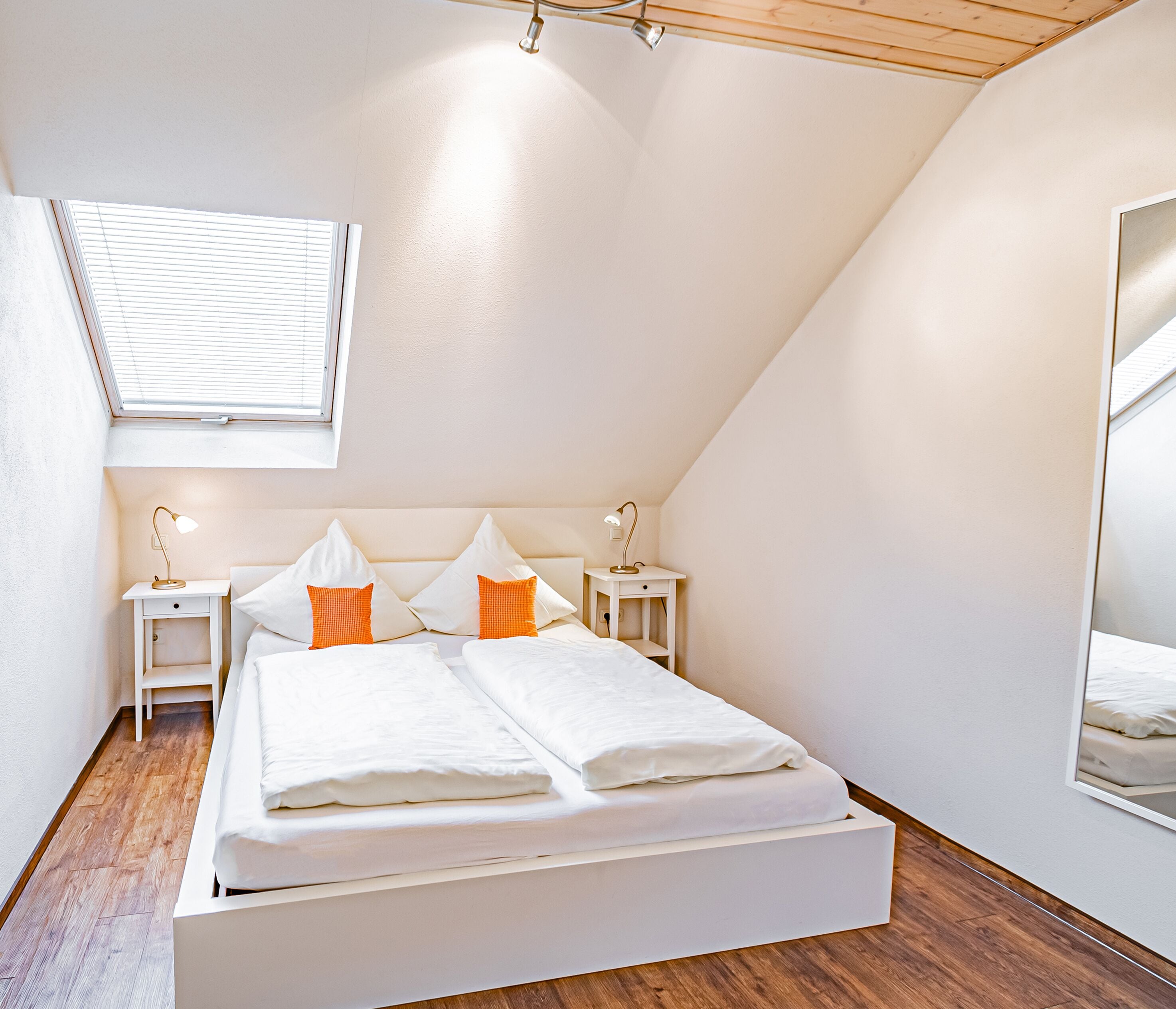 Um quarto moderno com um claraboia, uma cama com roupa de cama branca e almofadas laranja, duas mesas de cabeceira com abajures, e um espelho grande na parede.