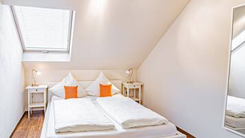 Um quarto moderno com um claraboia, uma cama com roupa de cama branca e almofadas laranja, duas mesas de cabeceira com abajures, e um espelho grande na parede.