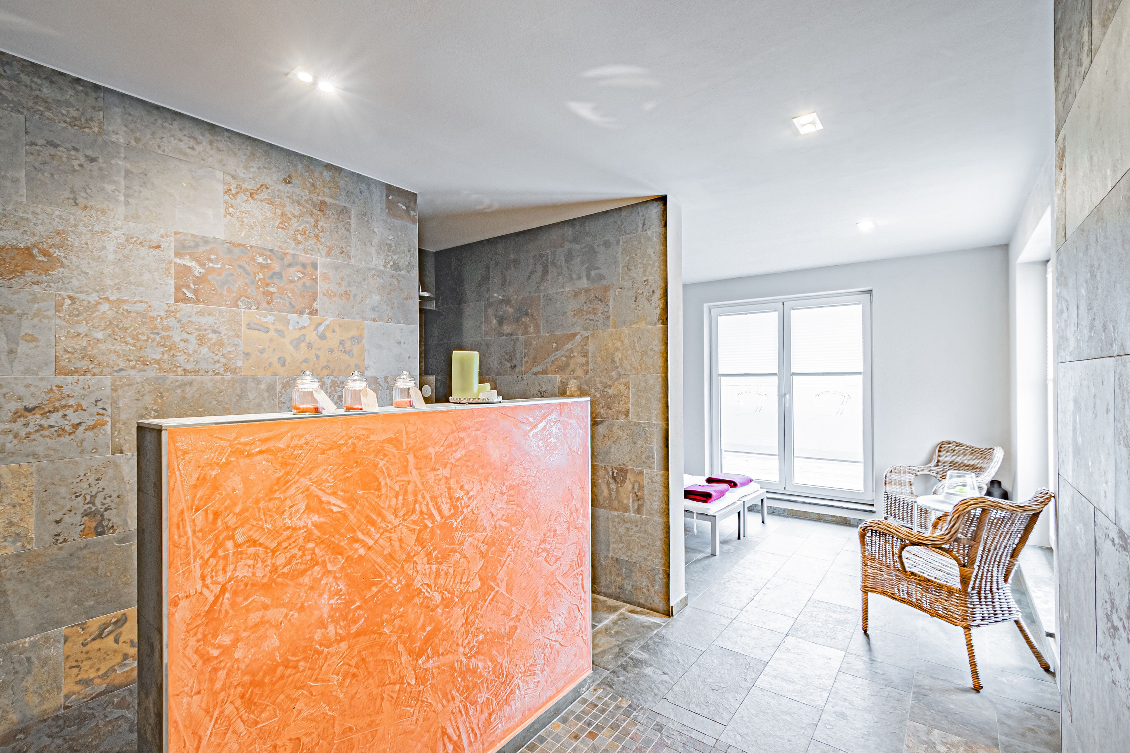 Un espace d’accueil avec un mur en pierre, un comptoir de réception, des fauteuils en osier et une grande fenêtre.