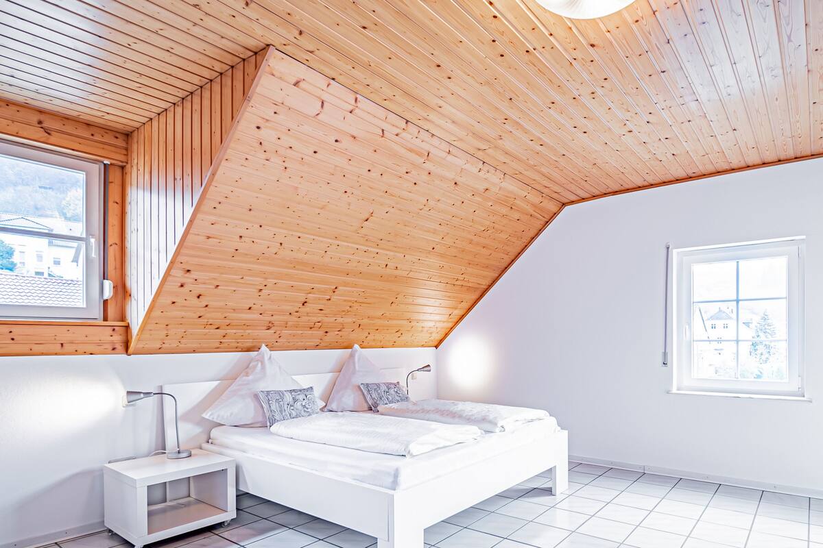 Une chambre avec un plafond en pente, des murs blancs, un lit, une table de chevet et une fenêtre.