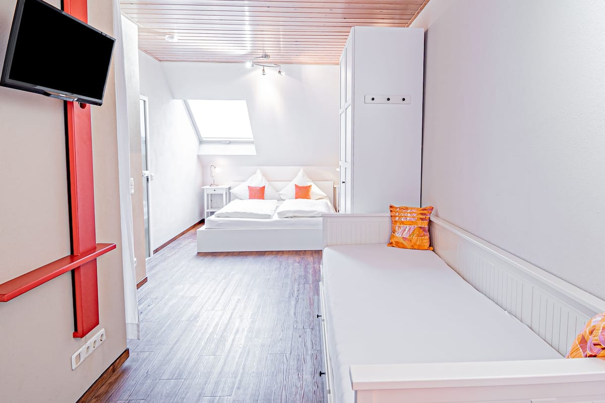 Une chambre moderne avec un plafond en pente, un lit avec une literie blanche et des oreillers orange, un banc blanc, un téléviseur fixé au mur et une lucarne.