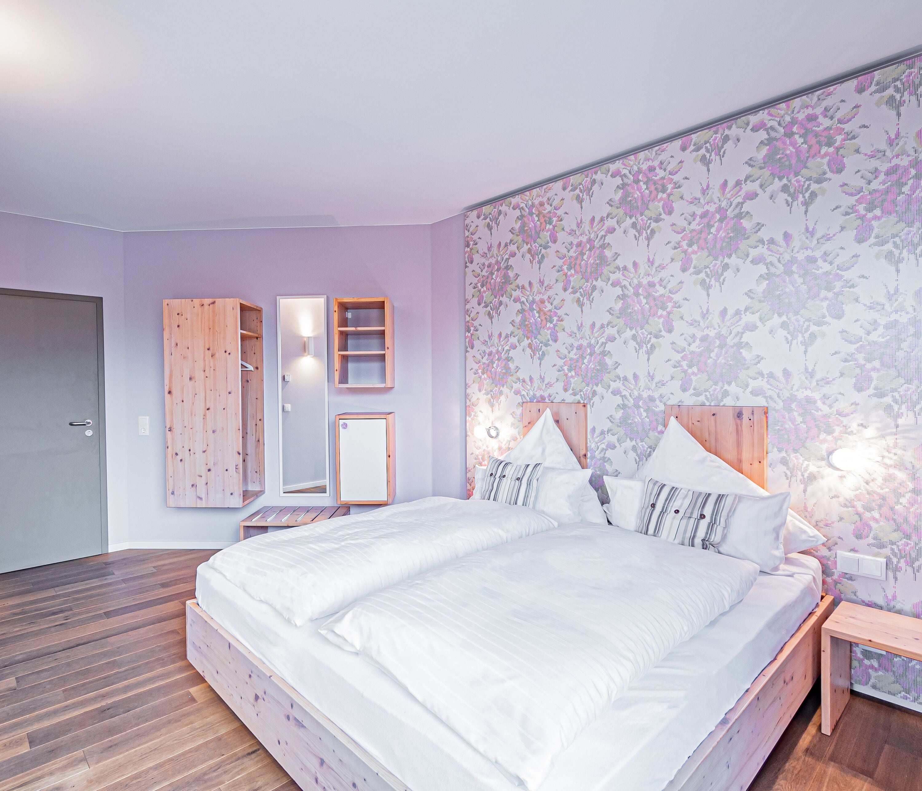 A bedroom with a large bed, wooden headboard, a nightstand, a mirror, and a wall-mounted shelf.