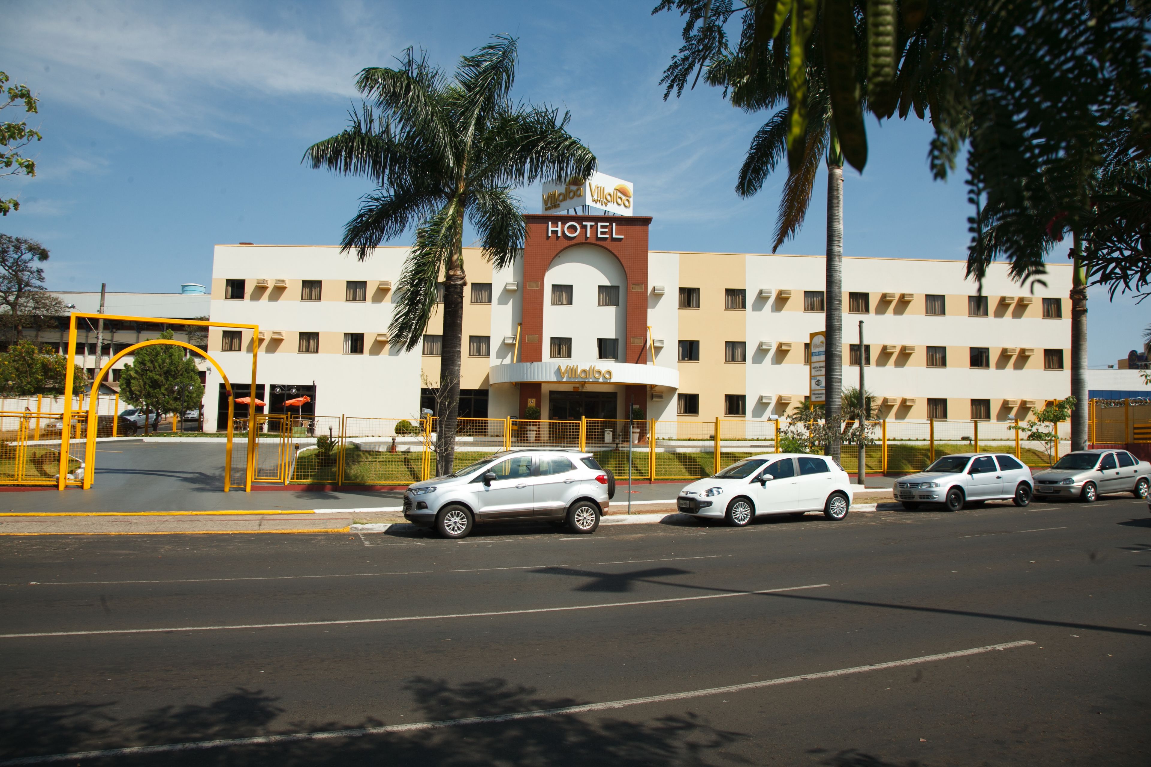 A hotel with a prominent sign, a parking lot with several cars, and palm trees in the foreground.