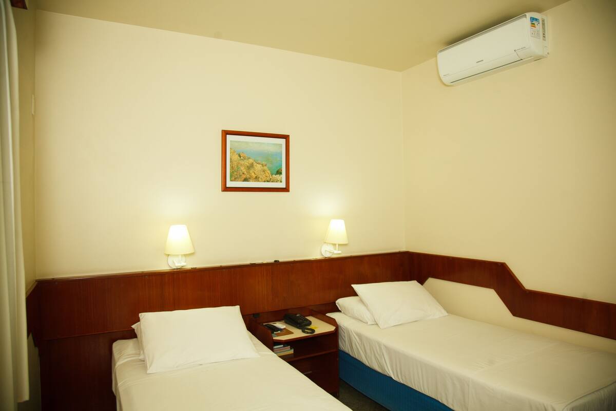 Une chambre d’hôtel avec deux lits simples, une table de chevet avec une lampe, un tableau encadré au mur et un appareil de climatisation.