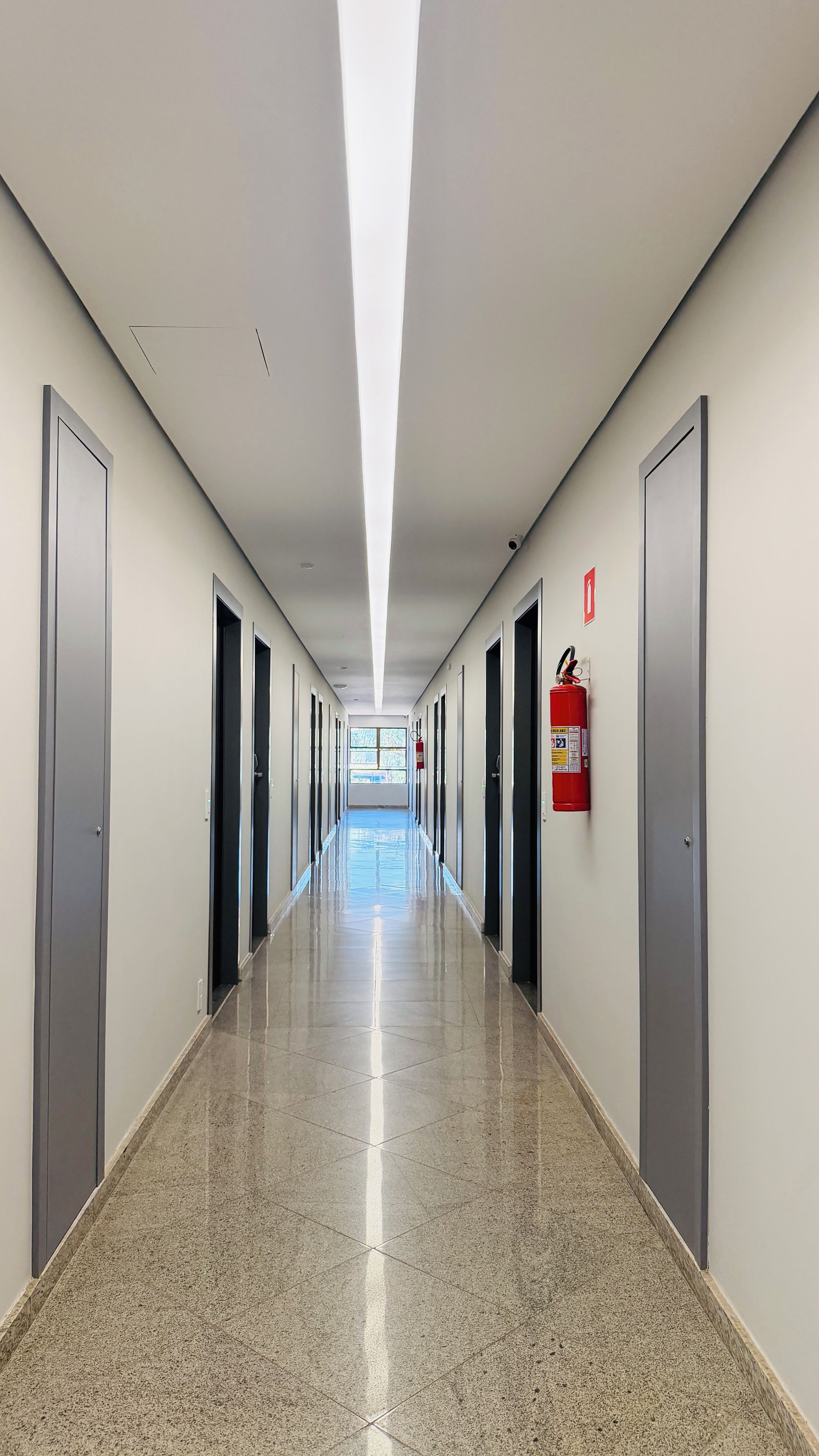 Um corredor longo e reto com portas em ambos os lados, um extintor de incêndio fixado na parede e uma janela no final.