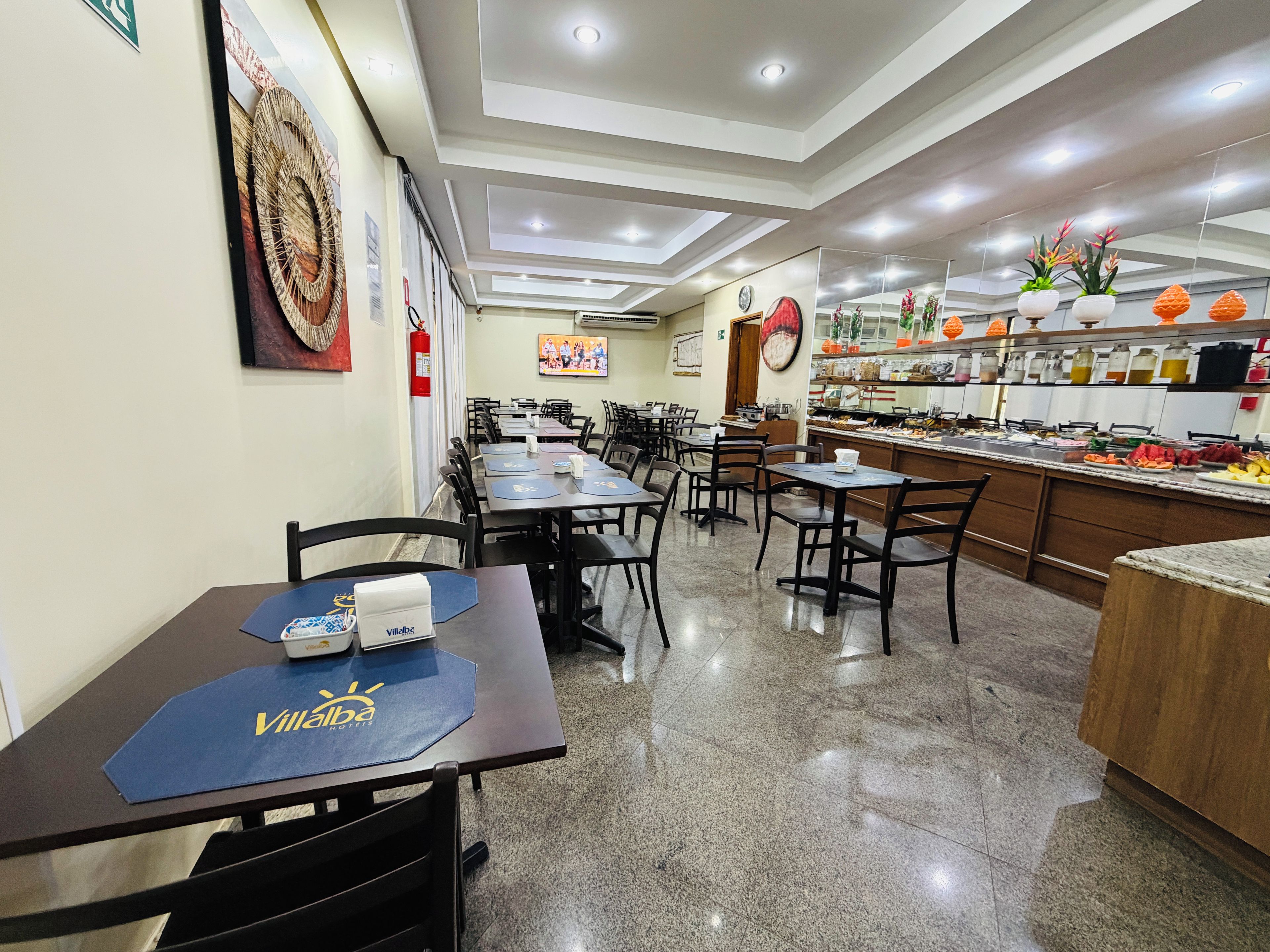 Un comedor de hotel con varias mesas y sillas, una barra de cocina con distintos alimentos, y un extintor en la pared.